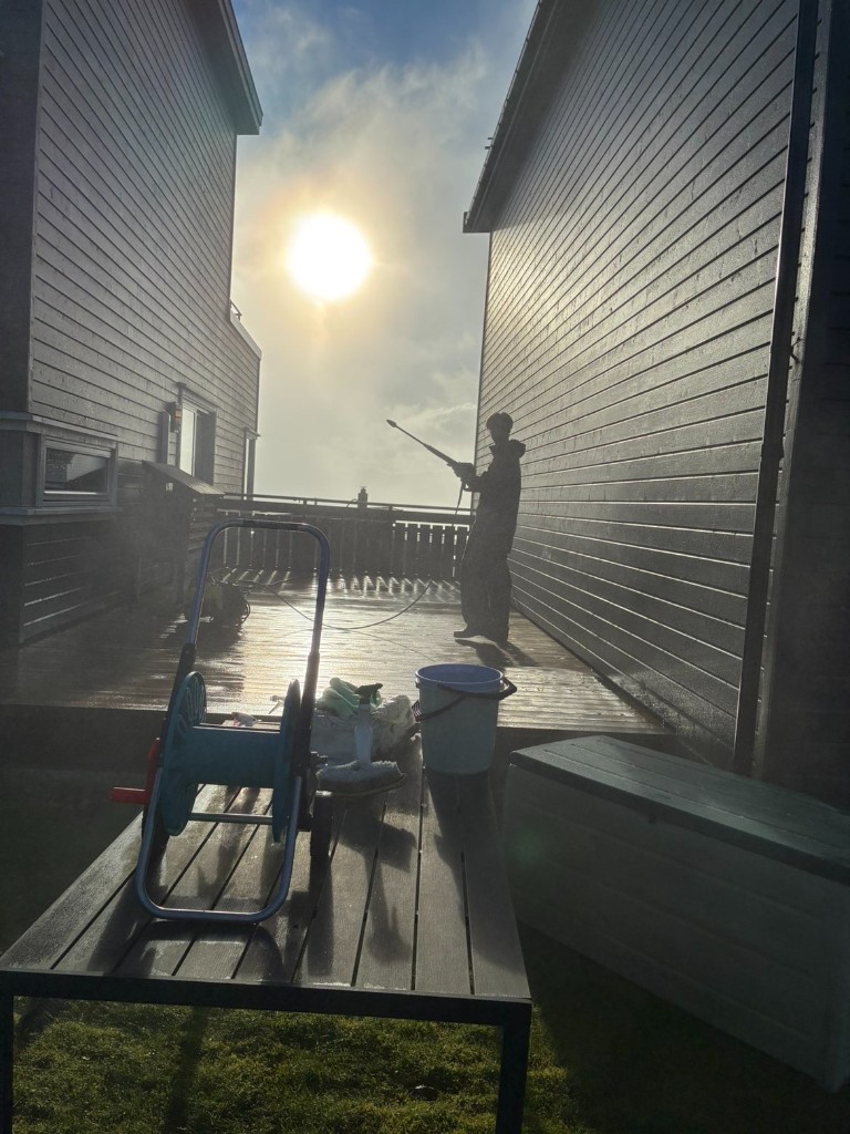 En av Hjelperne vasker en terrasse med høytrykksspyler i motlys fra sola.